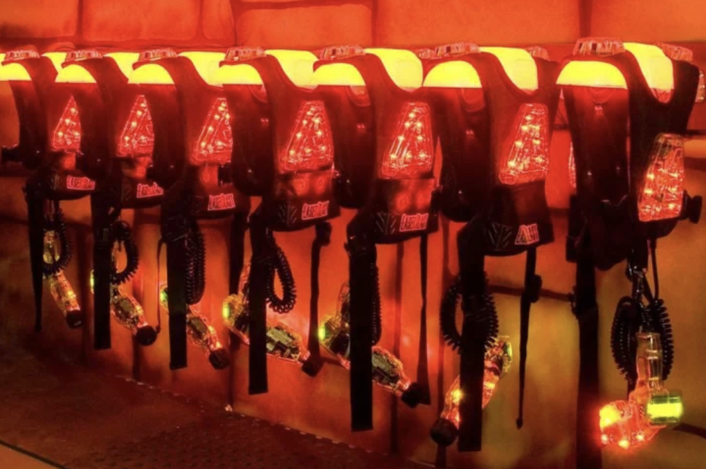 A row of illuminated laser tag vests and guns hang on a wall, ready for players. The equipment glows with red and orange lights, reflecting a futuristic, action-packed atmosphere in a dimly lit room.