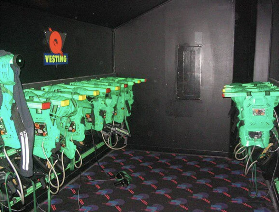 A dimly lit room with green laser tag vests and phasers hanging on black walls. The vests are lined up neatly, with wires connected to a central system. The floor is covered with a black carpet featuring colorful patterns.