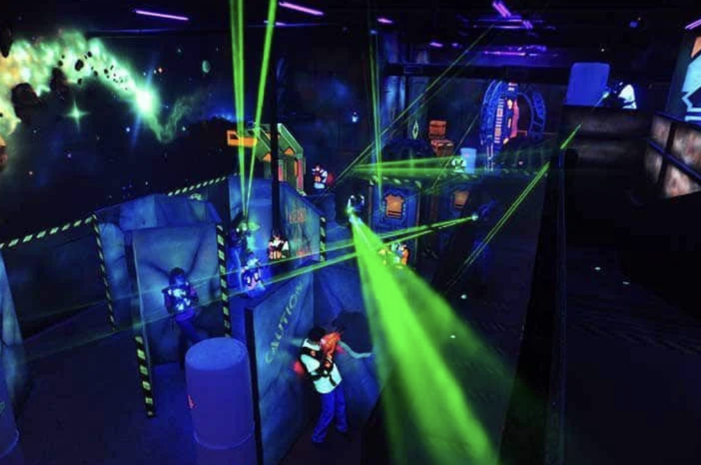 People playing laser tag in a dark arena with vibrant green laser beams and glowing walls decorated with space-themed artwork. Participants wear vests and use laser guns while navigating through barriers and obstacles.