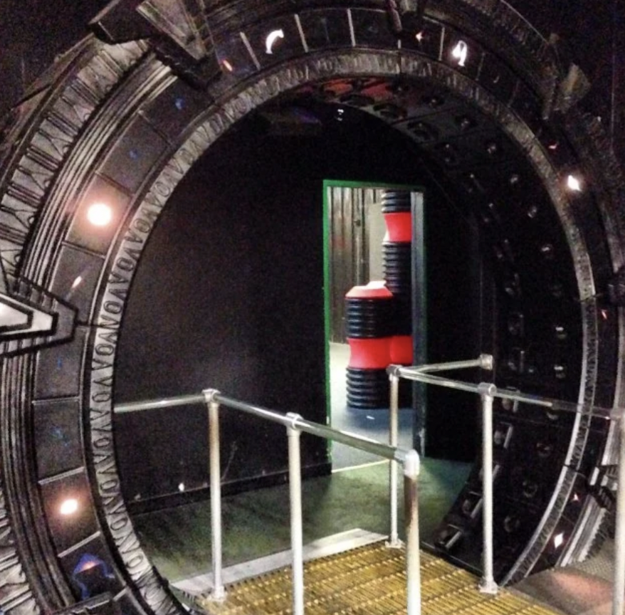 A large, circular, metallic structure resembling a sci-fi gateway, with glowing symbols around the edge, stands in a dimly lit room. A narrow ramp leads to its center. A doorway with red and black objects is visible in the background.