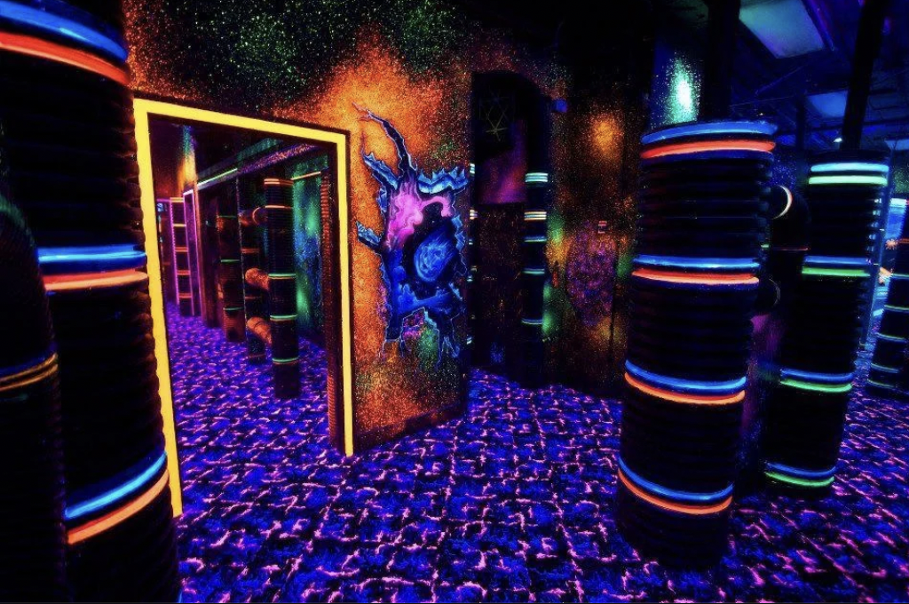 A vibrant, futuristic laser tag arena with glowing walls and neon-painted pillars. The room features blacklight designs, including a colorful cosmic mural. The floor is patterned with purple and blue geometric shapes, enhancing the otherworldly atmosphere.