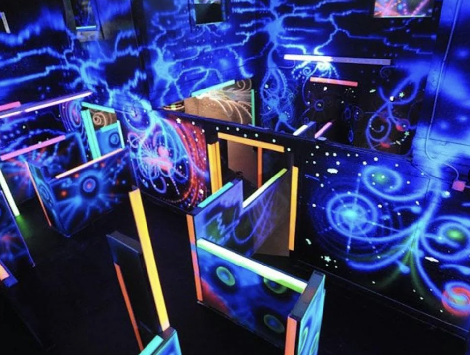 A vibrant laser tag arena with glowing neon walls and barriers. The walls display swirling blue, purple, and green designs with splashes of light creating a dynamic, energetic atmosphere. Brightly lit pathways create a maze-like layout.