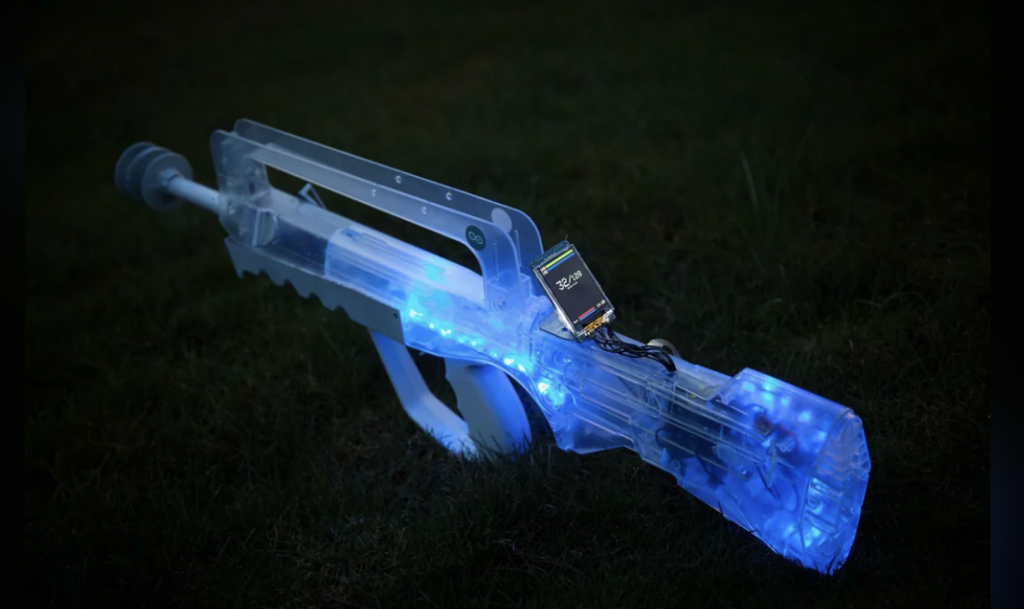 A transparent plastic gun illuminated with blue LED lights rests on grass in a dark setting. A small electronic device is attached near the top, glowing with different colors.