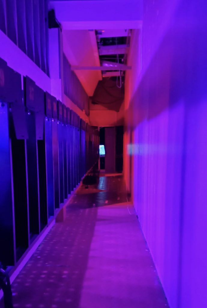 A narrow corridor illuminated by purple and pink lights, with tall lockers lining the left side. The floor is reflective, and in the distance, a backlit screen or door is visible. The scene has a futuristic, ambient feel.