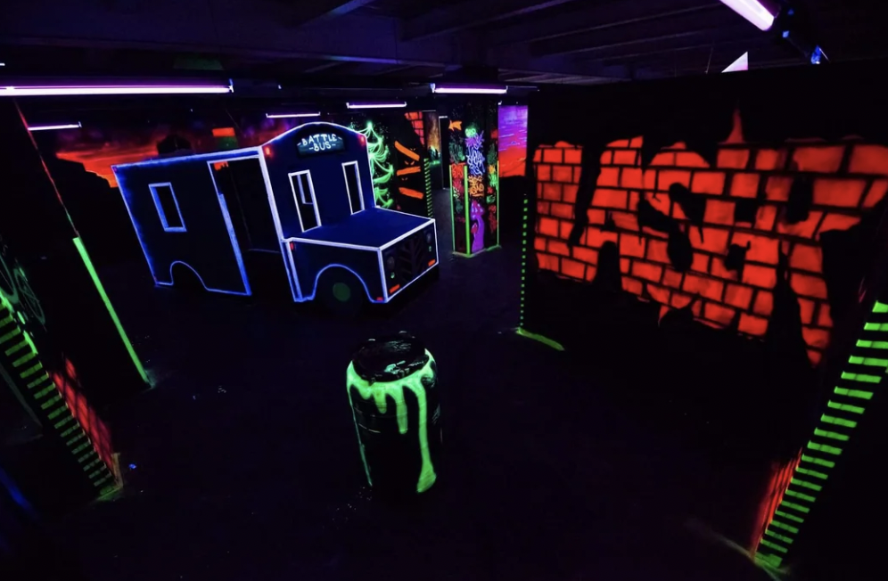 A dark, indoor laser tag arena with neon graffiti-style designs. Structures resemble urban buildings with vibrant colors, and a glowing barrel is in the center. The word "BLAZER" is spray-painted on a wall in bright letters. Purple lighting enhances the scene.