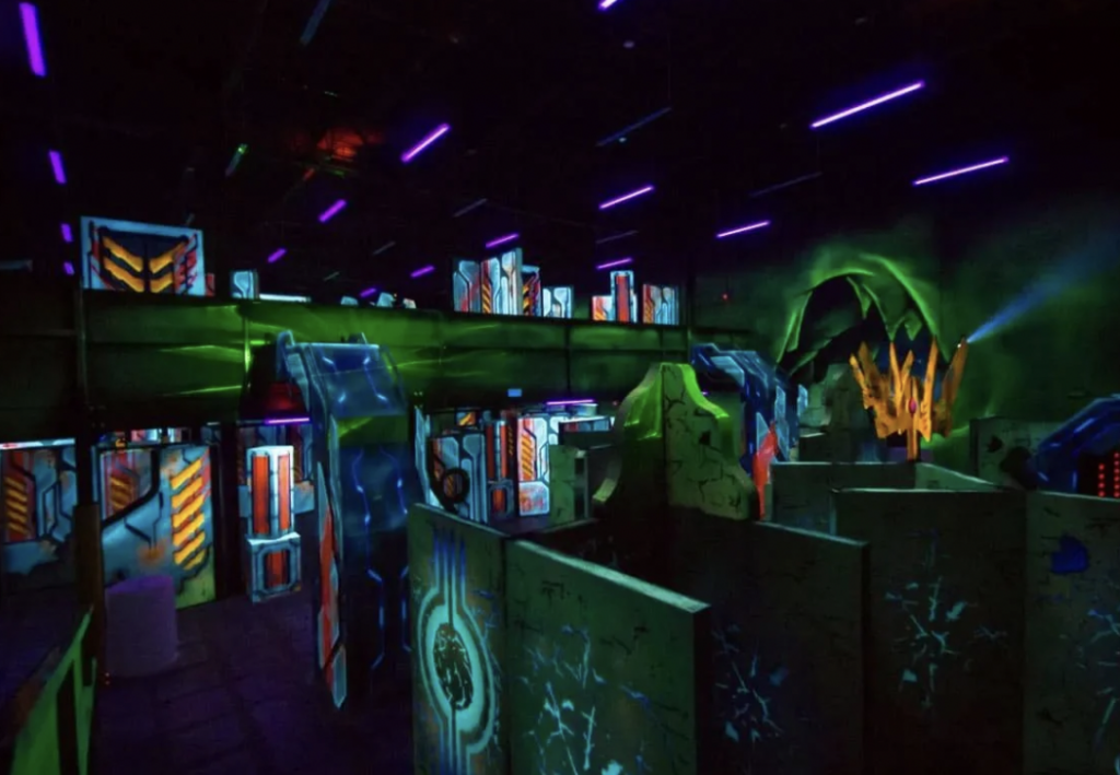 A vibrant laser tag arena with colorful neon lights and glowing obstacles. The space features various walls and barriers painted in bold patterns and designs, illuminated by purple and green lighting, creating an exciting and energetic atmosphere.