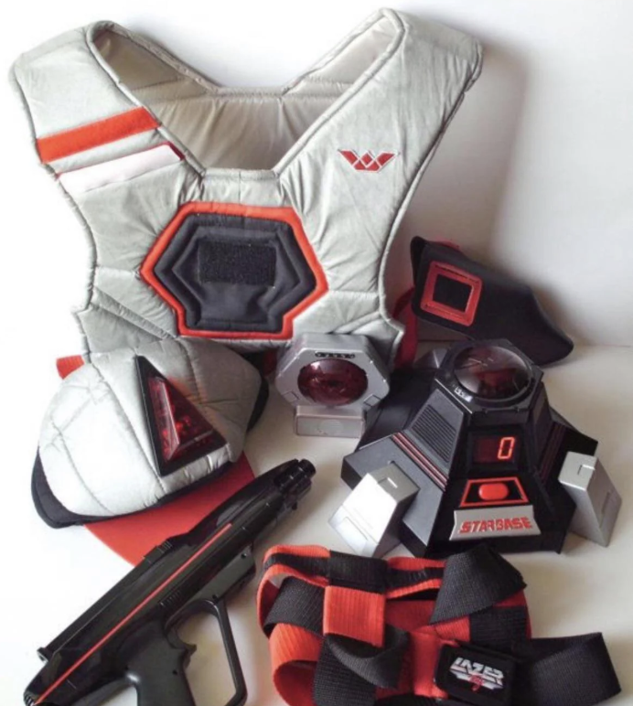 A laser tag set featuring a grey and red vest, a futuristic-looking gun, sensors, and other attachments laid out on a white surface. The equipment is detailed with various buttons and straps.