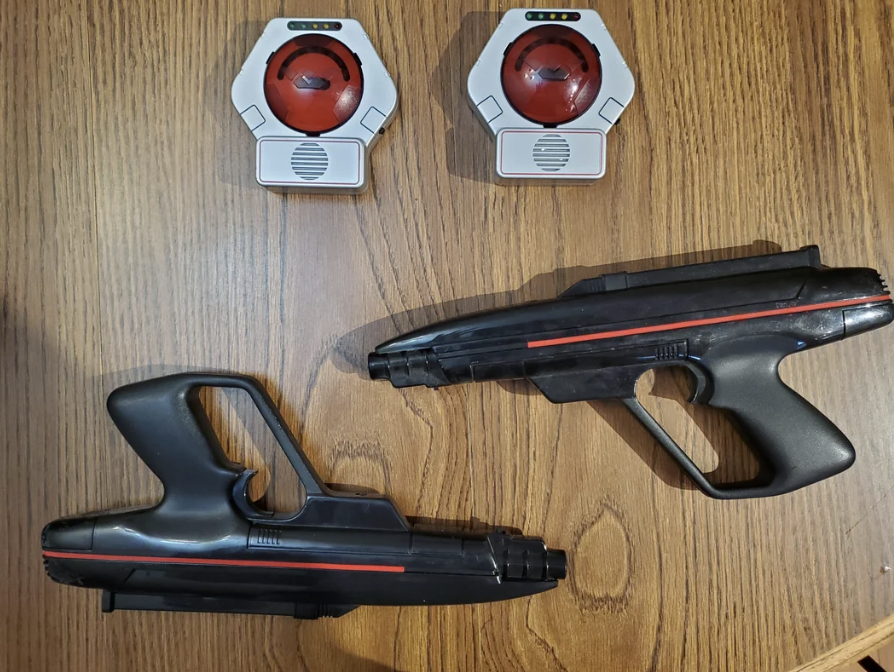 Two black and red toy laser guns are placed on a wooden surface alongside two round, red sensors in white holders.