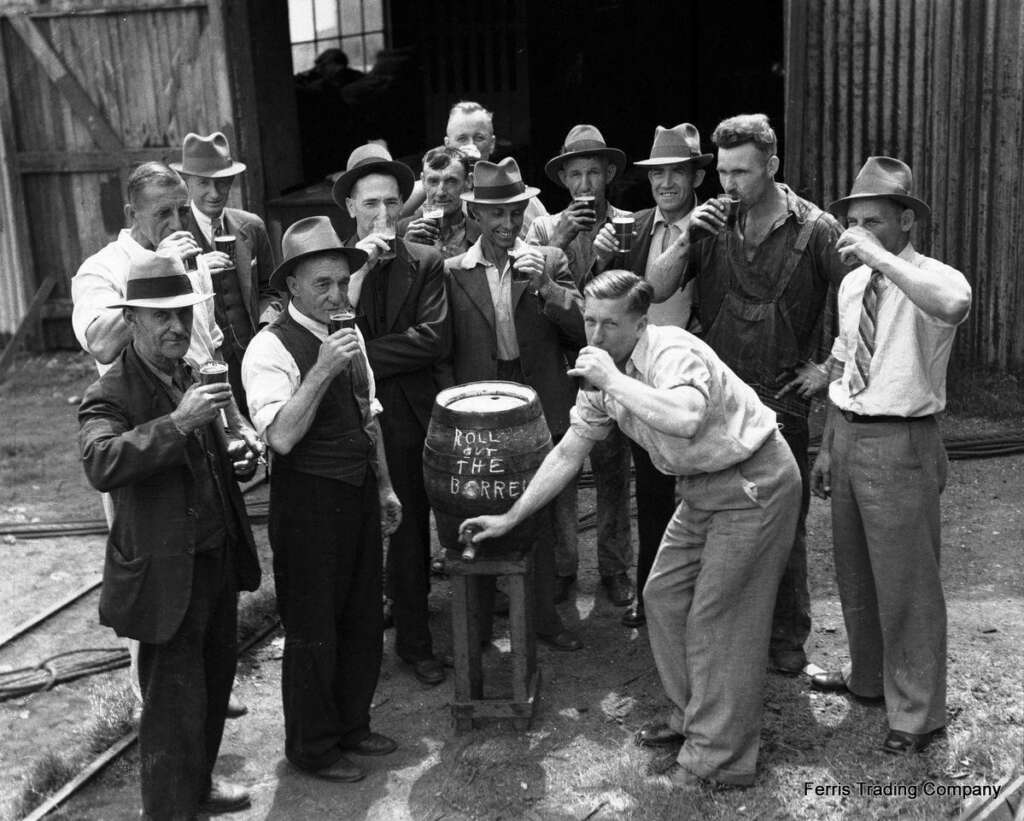 A group of men, dressed in vintage attire with hats, gather around a barrel labeled "Roll out the Barrel." Most are drinking from mugs. The setting appears to be an outdoor area with a wooden structure in the background.