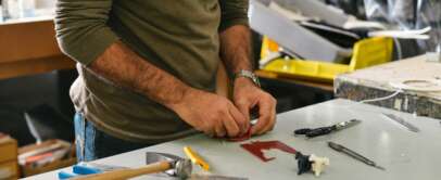 A person is working with tools on a table. Various hand tools, including a hammer and screwdrivers, are scattered around. The person is focused on a small piece of material, possibly for a craft or repair project.