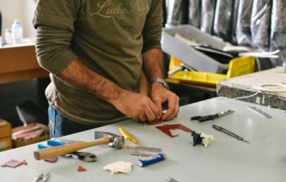 A person is working with tools on a table. Various hand tools, including a hammer and screwdrivers, are scattered around. The person is focused on a small piece of material, possibly for a craft or repair project.