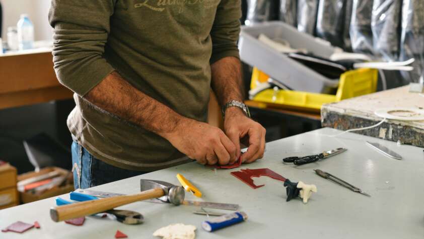 A person is working with tools on a table. Various hand tools, including a hammer and screwdrivers, are scattered around. The person is focused on a small piece of material, possibly for a craft or repair project.