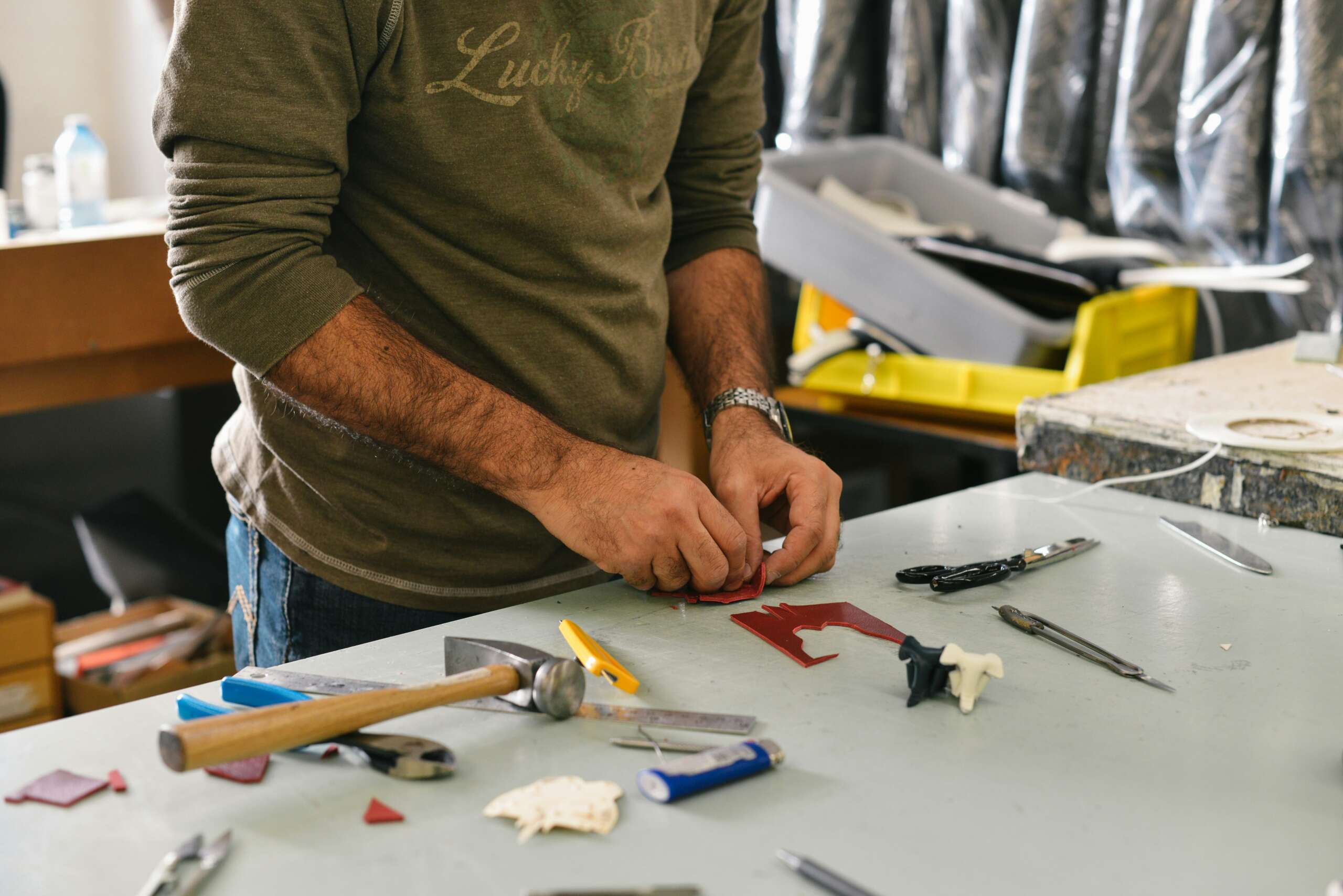 A person is working with tools on a table. Various hand tools, including a hammer and screwdrivers, are scattered around. The person is focused on a small piece of material, possibly for a craft or repair project.