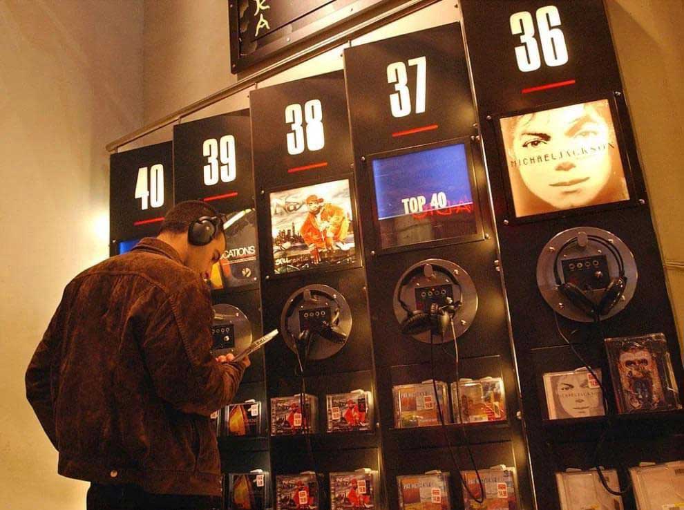 A person wearing headphones is selecting music at a listening station in a store. The wall displays various albums numbered 36 to 40, with CDs and promotional images visible. The ambiance appears warm and inviting.