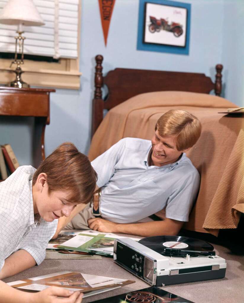 Two people are in a room listening to vinyl records. One is sitting on the floor, leaning against a bed, while the other is lying on the floor. A record player and albums are in front of them. The room has a bed, lamp, and posters on the walls.