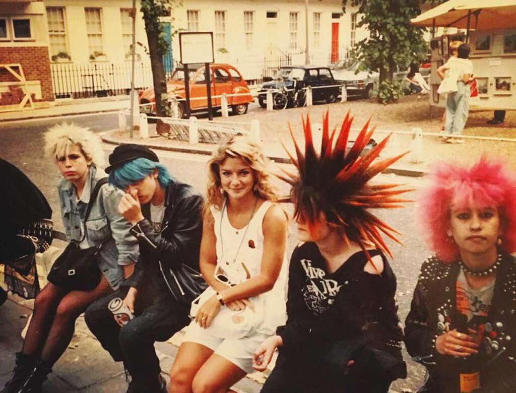 A group of five people sitting on a street bench. Four have colorful punk hairstyles and attire. The central person, with blond hair, is smiling. In the background, vintage cars and a street corner are visible. The scene has a retro feel.