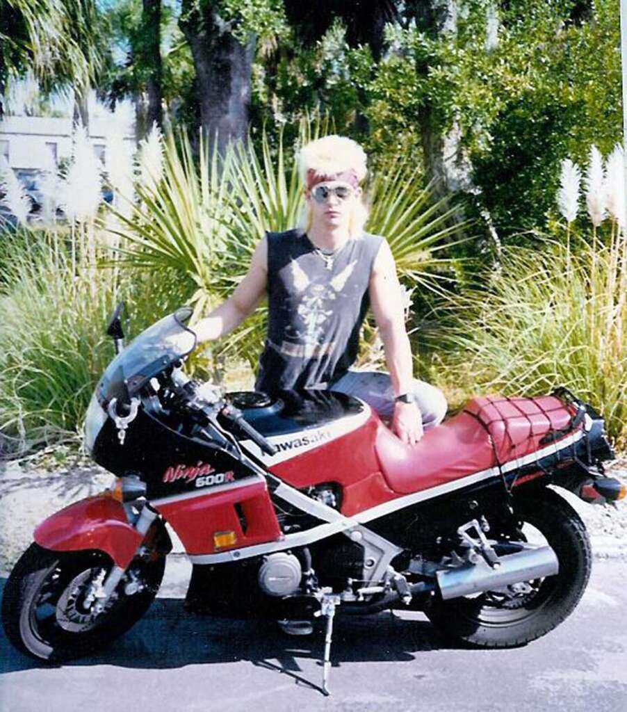 Person with blonde hair and sunglasses sits on a red Kawasaki Ninja 600R motorcycle. They are wearing a sleeveless shirt. Green foliage and trees are in the background, with a sunny atmosphere.