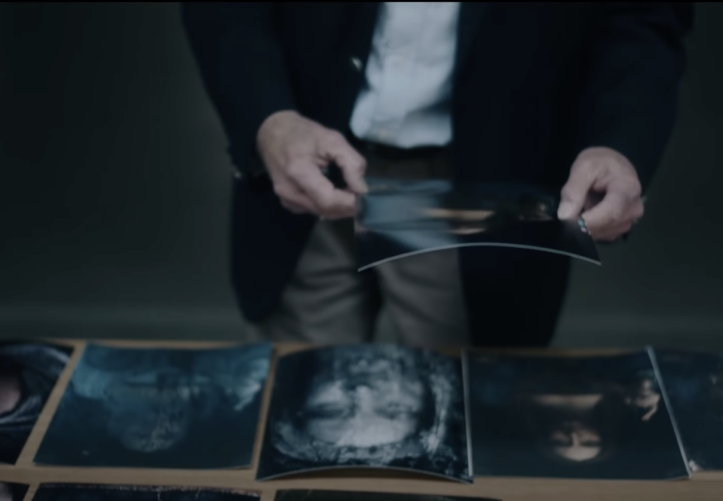 A person in a dark jacket and light pants holding a photo above a table. The table is covered with various photographs, some featuring faces, laid out against a dark background.