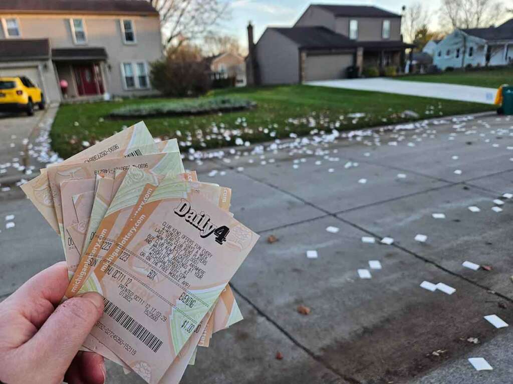 A hand holding lottery tickets is in focus in the foreground. In the background, a neighborhood street is littered with white paper scattered on driveways and lawns. Houses and a yellow car are visible.