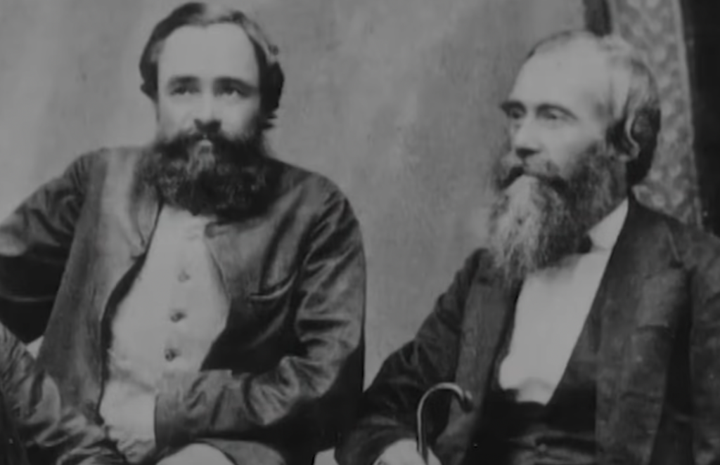 Two bearded men in vintage attire are seated next to each other. The man on the left wears a buttoned jacket, while the man on the right wears a suit and holds a cane. The backdrop is simple and textured.