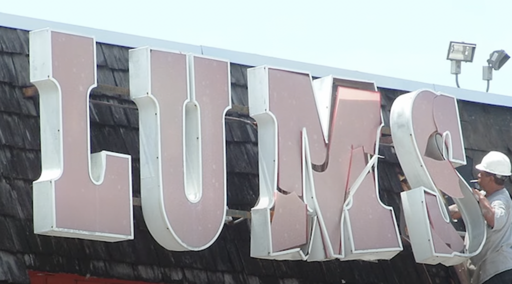 A worker on a ladder removes a large, red and white "S" from a sign that reads "LUMS" on a building with a shingle roof. The sky is clear in the background.