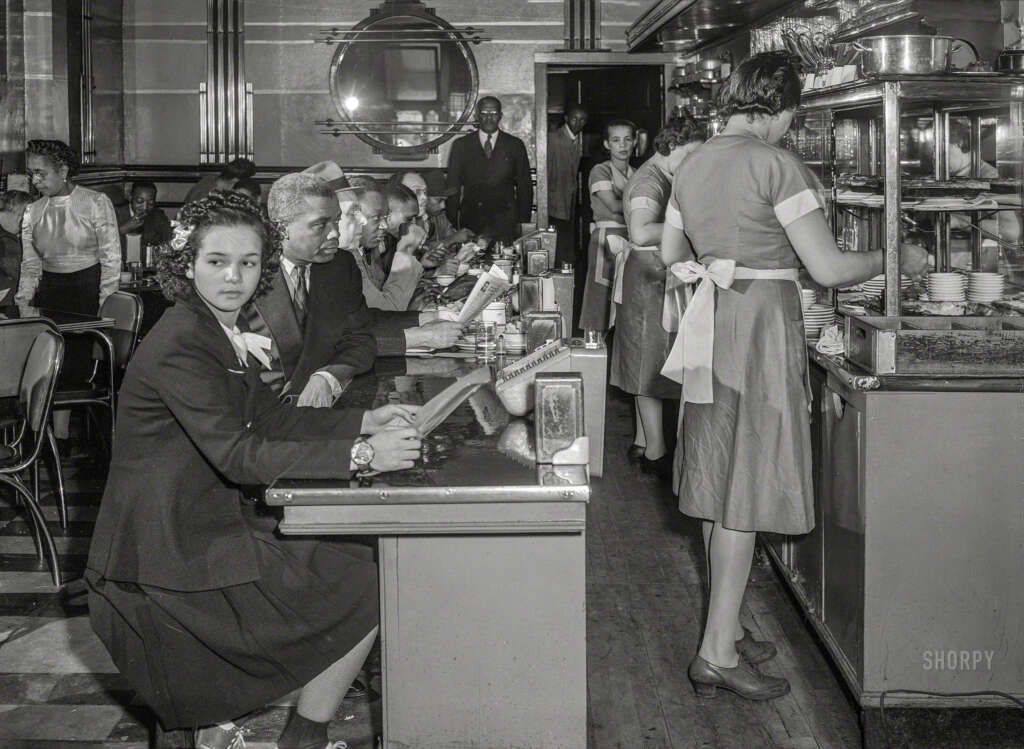 Black and white photo of a busy 1940s diner. A woman sits at a counter on the left, wearing a dark suit and white blouse. Waitresses in uniforms serve customers. The diner has checkered floors and reflective surfaces. People are eating and conversing.