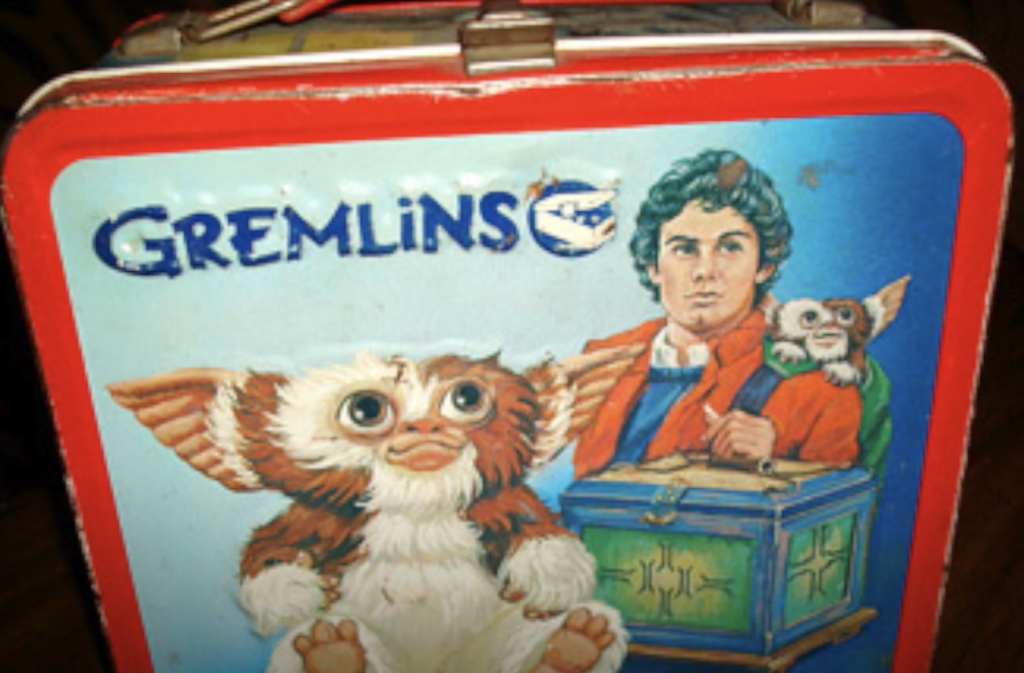 A vintage metal lunchbox featuring "Gremlins" artwork. It shows a character holding a small creature on their shoulder, with a larger creature sitting below. The background is blue, and the word "Gremlins" is at the top.