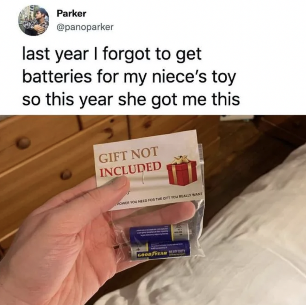 A hand holds a small package containing AA batteries labeled "Gift Not Included." An insert reads "POWER YOU NEED FOR THE GIFT YOU REALLY WANT." The image includes a tweet saying, "last year I forgot to get batteries for my niece’s toy so this year she got me this.