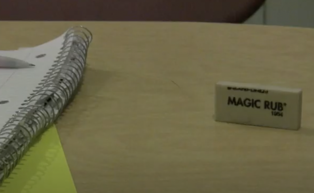 A close-up of a desk with a spiral notebook, a pen, and a Magic Rub eraser. The notebook is open, resting on a paper with a yellow sheet beneath it. The items are on a wooden surface.