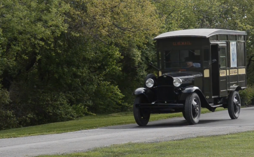 A vintage U.S. Mail truck from the early 20th century drives on a paved road. The vehicle is dark green with a white roof and large round headlights, surrounded by lush green trees and grass.