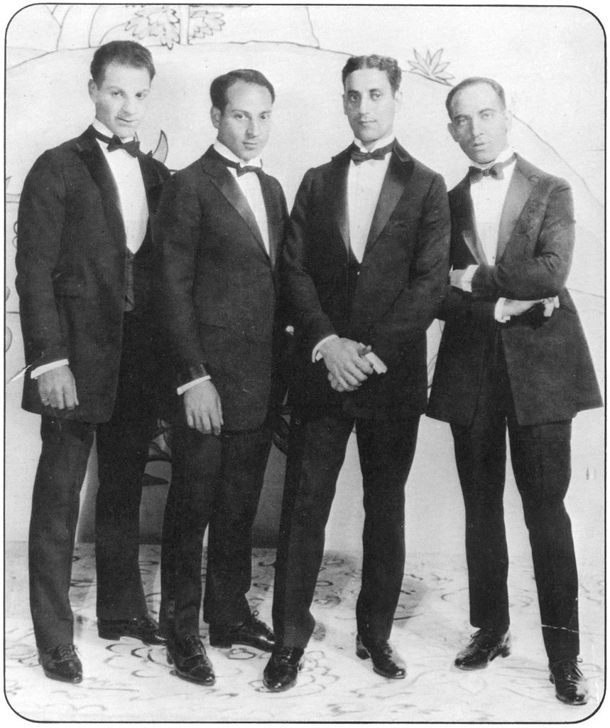 Four men in tuxedos stand side by side against a painted backdrop with floral and abstract patterns. They have slicked-back hair and formal poses, reminiscent of early 20th-century fashion.