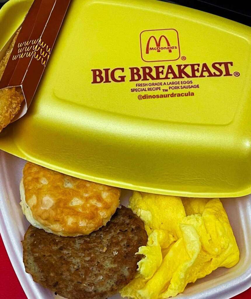 Yellow McDonald's Big Breakfast container open, revealing scrambled eggs, a sausage patty, and a biscuit. A hash brown partially visible next to the container.