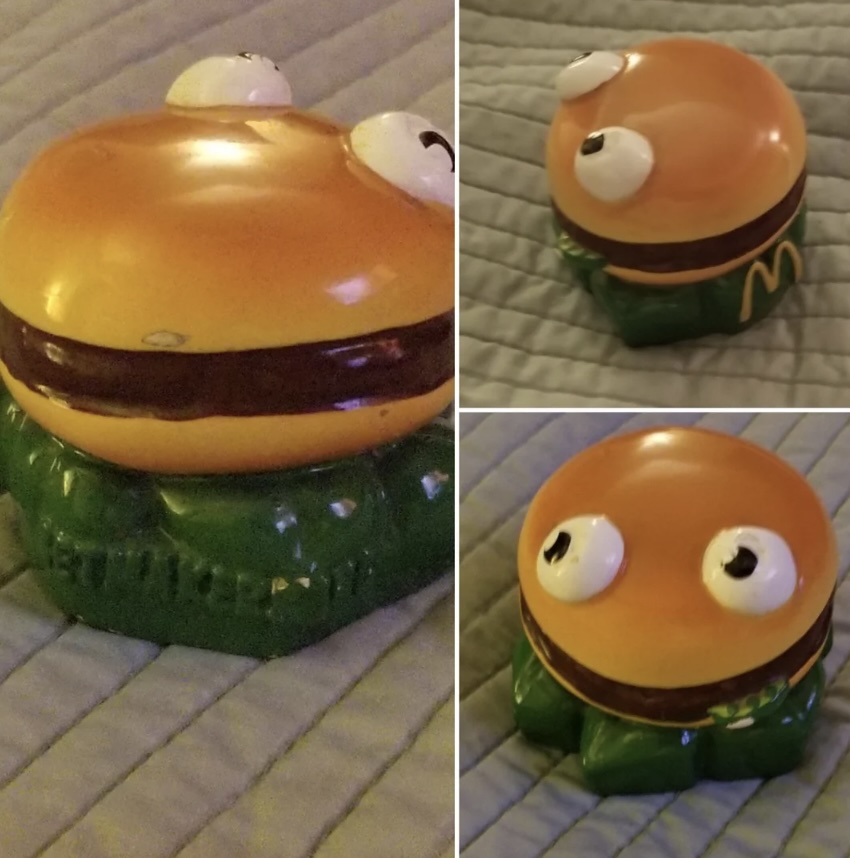 A ceramic figure resembling a burger with eyes sits on a quilted gray surface. The burger appears with a bun on top and bottom, white eyes, and a green base. Three angles are shown: front left, back right, and top view.