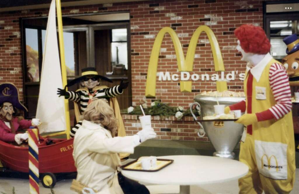 A whimsical McDonald's scene featuring characters in vibrant costumes. Ronald McDonald serves food at an outdoor table. The Hamburglar waves, standing next to a brick facade with the McDonald's logo. A boat with a captain character is on the left.