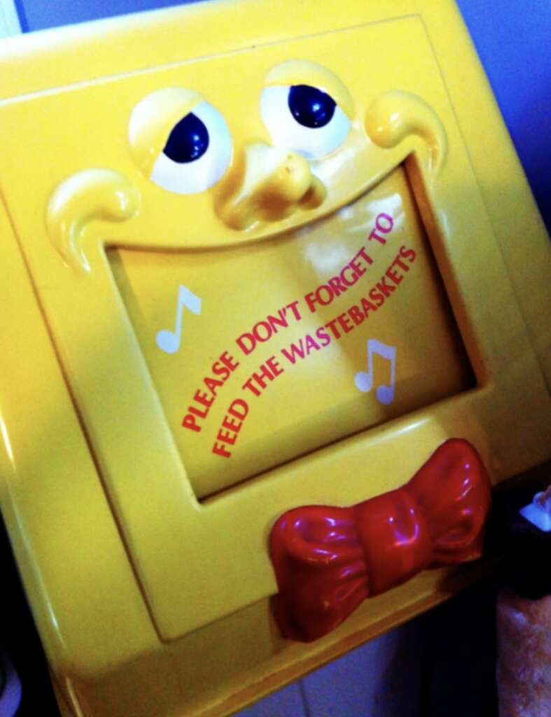 A yellow wastebasket with a smiling face and a red bow tie. The flap reads, "PLEASE DON'T FORGET TO FEED THE WASTEBASKETS," with musical notes around the text.