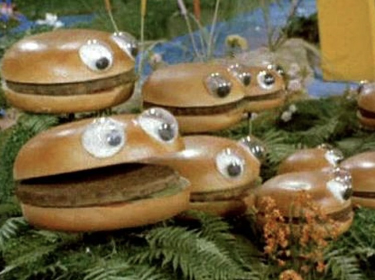 A group of anthropomorphic burgers with eyes and mouths sits on grass and ferns. The burgers appear animated with expressive eyes and different mouth shapes, giving them playful personalities.