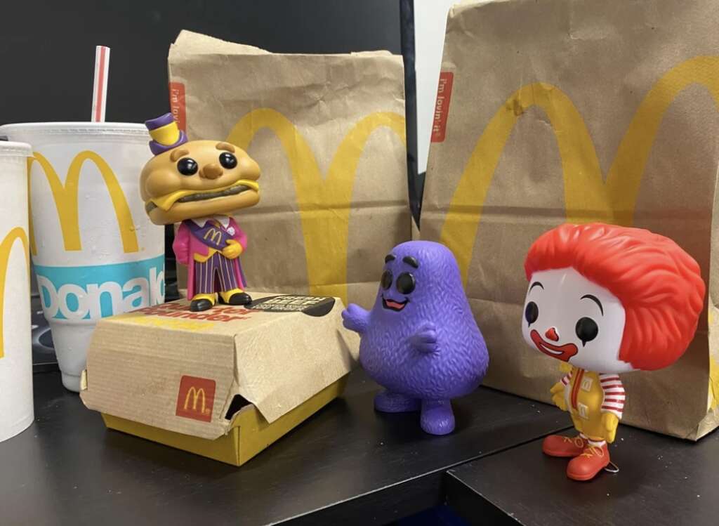 Three toy figures resembling classic fast-food characters are surrounded by McDonald's paper bags and cups. A burger character, a purple blob, and a clown figure stand near a boxed meal, set on a black table.