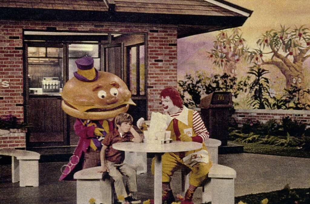 A child sits at an outdoor table with a character in a striped outfit and another character with a hamburger head wearing a purple hat and suit. They are enjoying a meal together in a garden-like setting near a brick building.