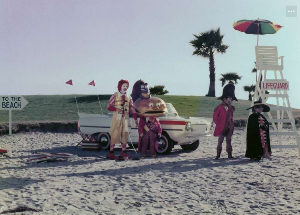 A group of whimsical costume characters, including a clown and others, stand on a sandy beach. A lifeguard chair with a colorful umbrella is nearby, and a small vintage car is parked in the background. Palm trees and a beach sign complete the scene.