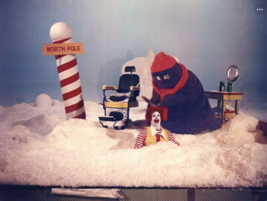 A colorful scene with a person in a clown costume sitting in fake snow at the North Pole, accompanied by a large purple creature wearing a red hat. A barber chair and mirror are nearby, with a candy-striped pole labeled "North Pole.