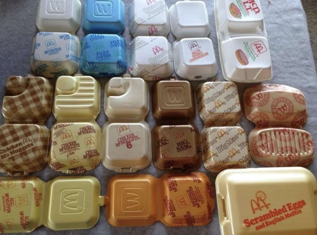 A collection of colorful vintage McDonald's containers, neatly arranged in rows on a grey surface. The containers display various designs and logos, predominantly featuring items like Big Mac, Quarter Pounder, and Egg McMuffin packaging.