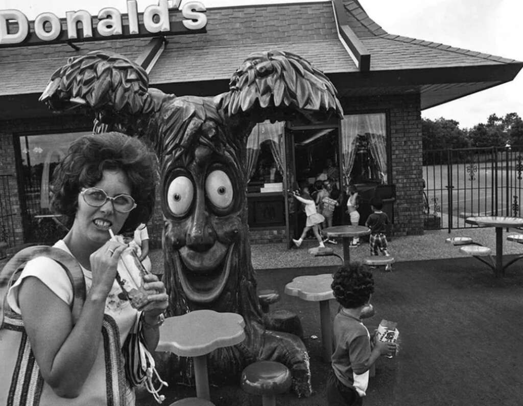 A woman with glasses and a bag eats outside a McDonald's, next to a tree-shaped sculpture with a surprised face. Children are entering the building in the background. The scene is in black and white.