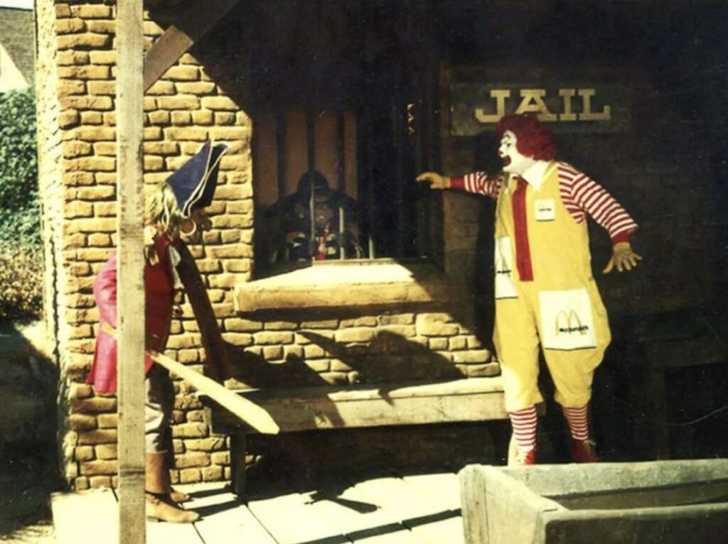 A scarecrow and a clown stand outside a jail with a person visible behind the bars. The clown wears a yellow suit with red stripes, and the scarecrow holds a wooden bat. The scene takes place in front of a brick building.