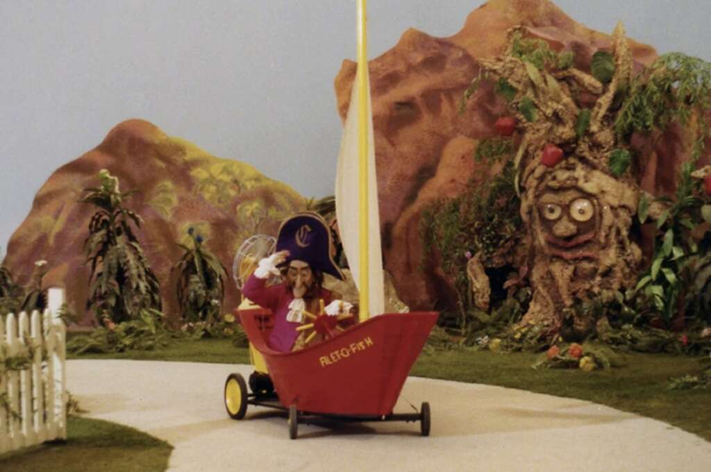 A whimsical scene with a puppet character dressed as a pirate, steering a red, boat-like vehicle labeled "Alco-Fish" on a white road. Surrounding are lush plants and a large animated tree with a face, set in a vibrant, colorful landscape.