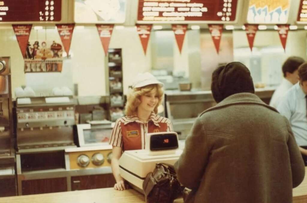 A fast-food restaurant scene from the 1970s or 1980s shows a cashier wearing a striped uniform and cap, smiling at a customer across the counter. The menu board in the background features classic burger options.