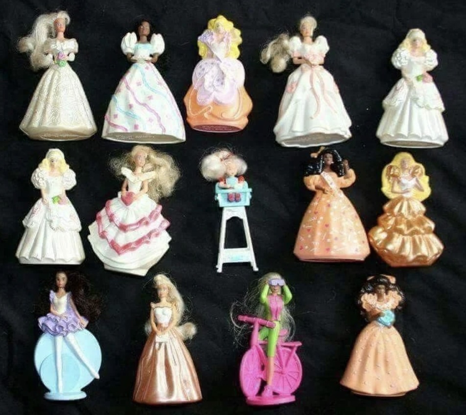 A collection of twelve doll-shaped containers with removable skirts arranged in rows. They feature various themes, colors, and styles, including a doll with a kitchen stove and one on a pink bicycle. All dolls have long hair and detailed dresses.