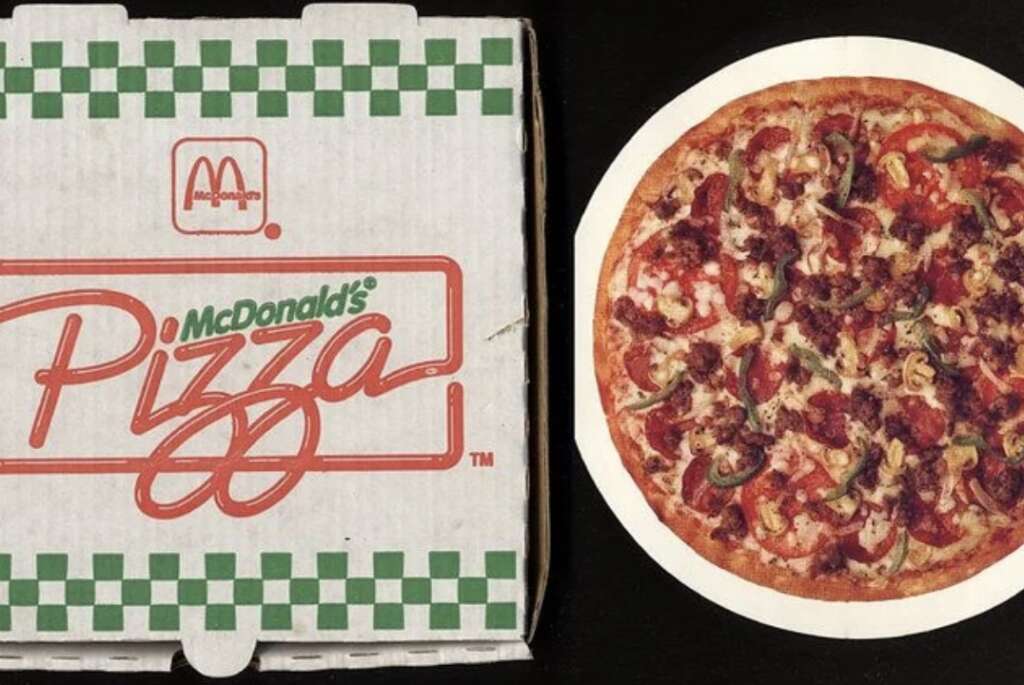 Image of a McDonald's-branded pizza box with a green and white checkered design next to a pepperoni and vegetable pizza on a black background. The word "Pizza" is prominently displayed on the box.