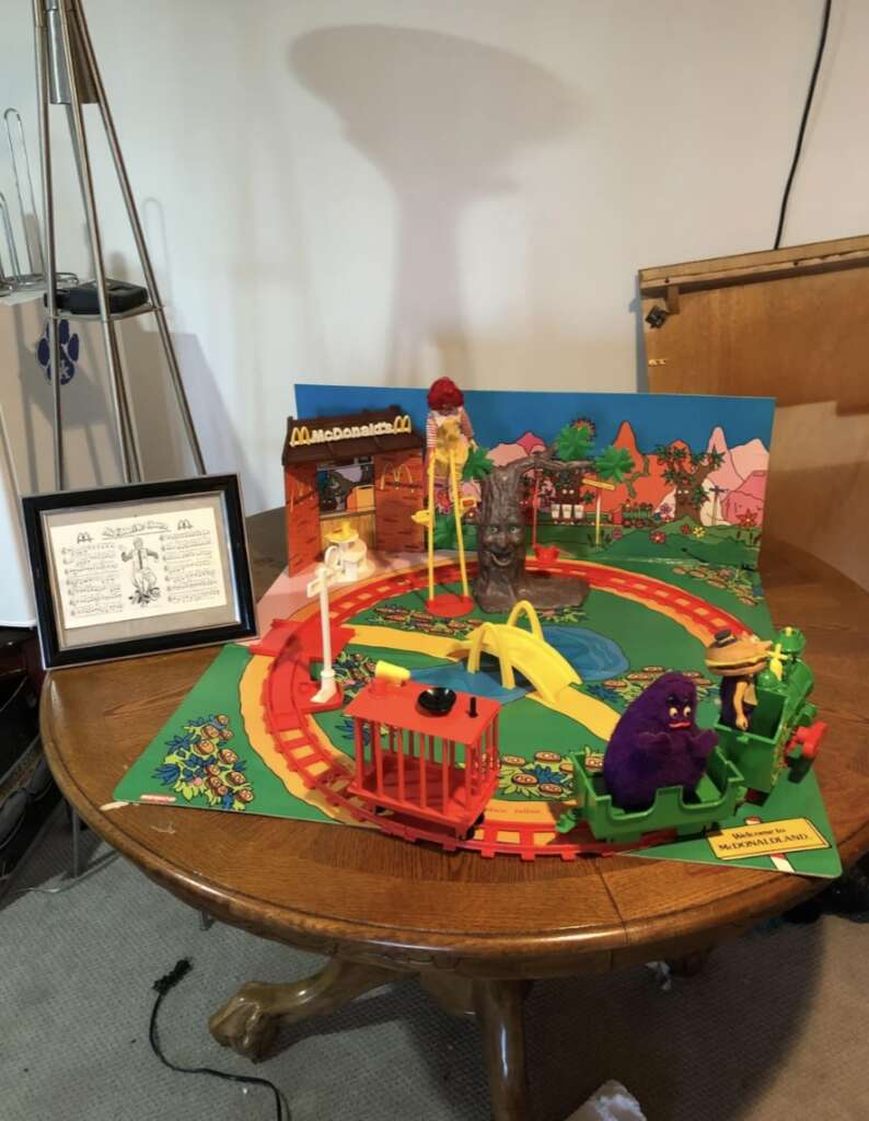 A display of McDonald's memorabilia featuring a colorful playset with various characters on a train track. The setup includes a model restaurant, trees, and a pond. A framed certificate stands on a wooden table beside the playset.