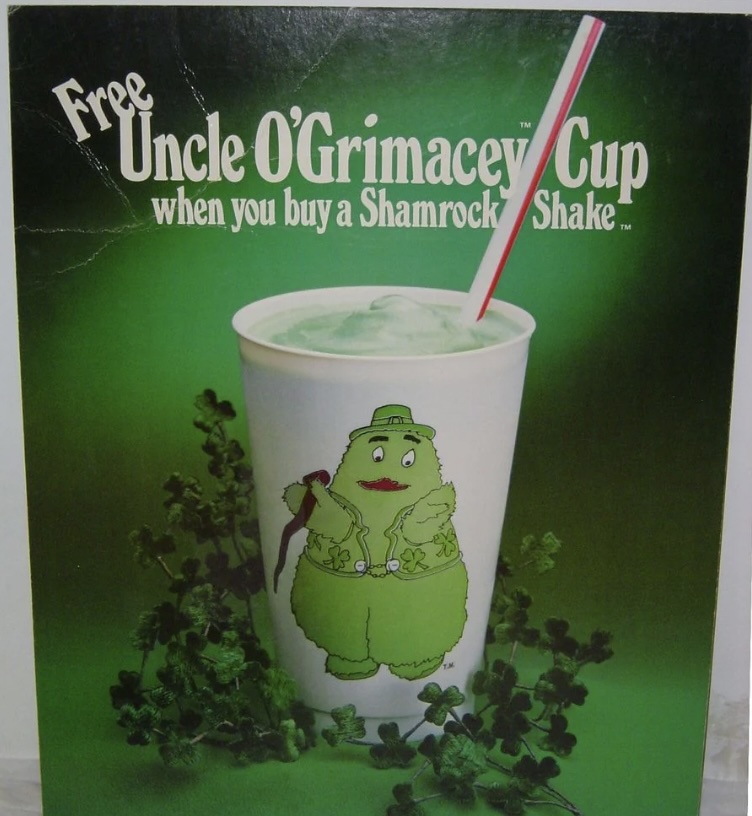 A promotional poster featuring a cartoon character named Uncle O'Grimacey on a cup, surrounded by clovers. The text reads "Free Uncle O'Grimacey Cup when you buy a Shamrock Shake." The background is green.