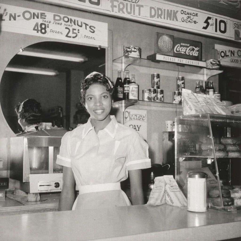 Black and white photo of a woman in a uniform standing behind a counter in a diner. Signs above list prices for donuts and drinks. A mirror reflects part of the counter. Various items are displayed, including a bottle and packaged goods.